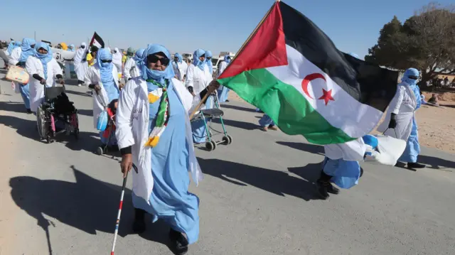 Une femme âgée portant un foulard bleu et blanc porte un drapeau du Sahara occidental