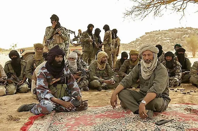 Combattants touaregs et arabes réunis sous un arbre, discutant autour d'un tapis traditionnel.